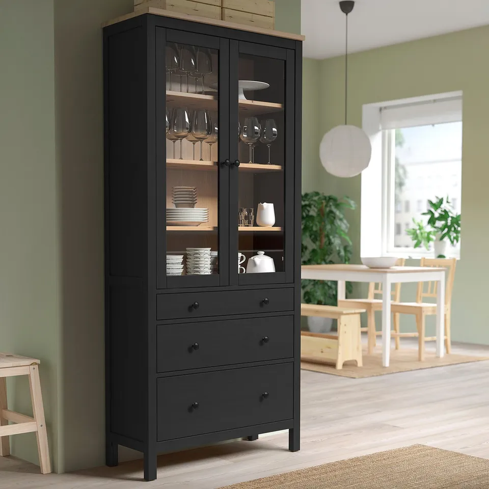Black HEMNES cabinet with glass doors, wooden shelves, and glass/dishware. Tall near green wall.
