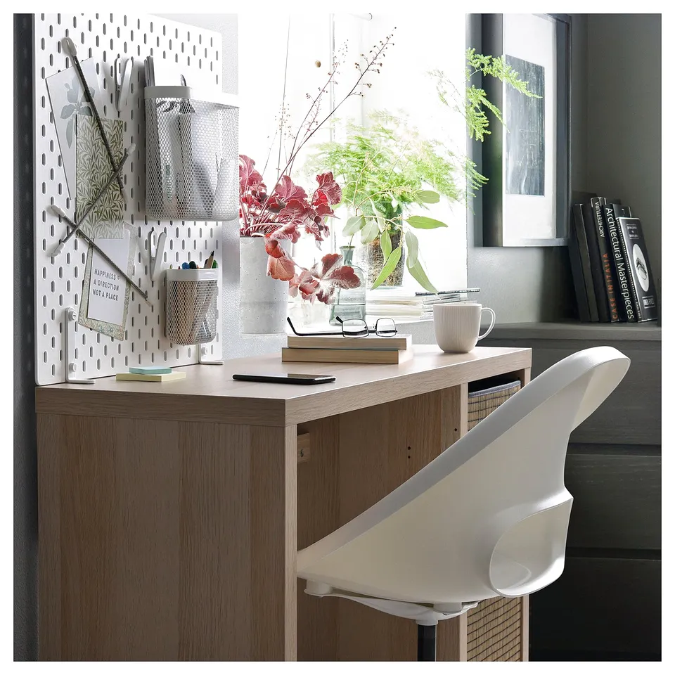 Desk: KALLAX in light brown, wooden, rectangular, with white chair; wall pegboard, flowers, cup, books.
