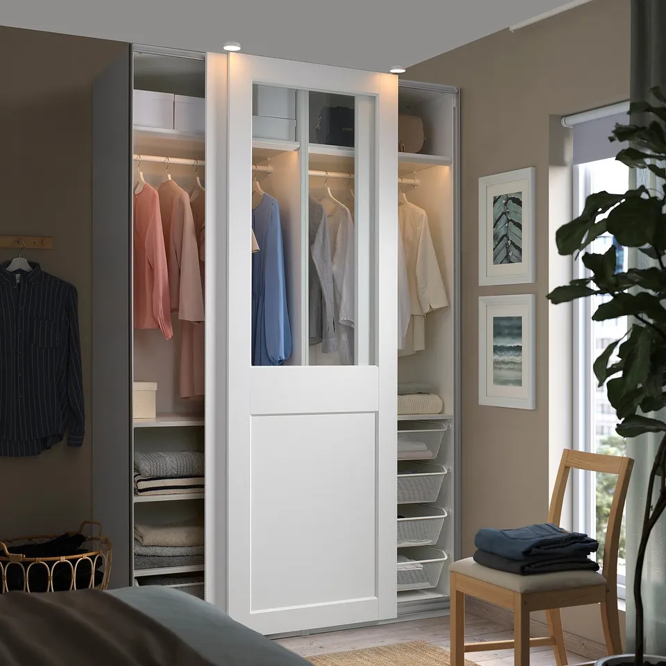 Modern white PAX wardrobe, half glass, holding clothes and shelves. Tall, fits in room corner, near large window.