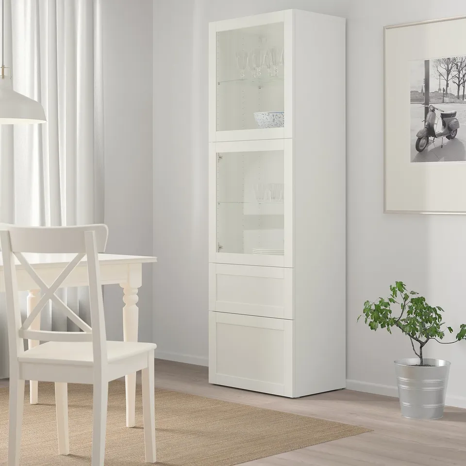 White BESTÅ cabinet with glass doors displaying wine glasses and dishes. It stands in a bright room next to a white chair, two drawers, and is taller.