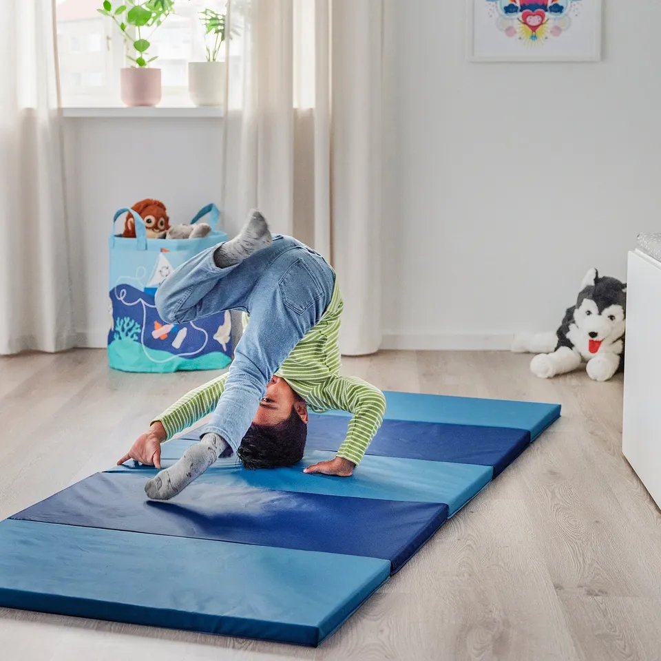 Person practises yoga on blue foldable mat in bright room, surrounded by toys.