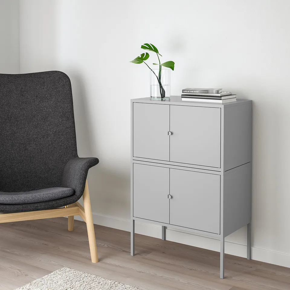 Grey LIXHULT cabinet with 2 doors, slim legs. Beside a dark chair, it holds a plant vase and books.