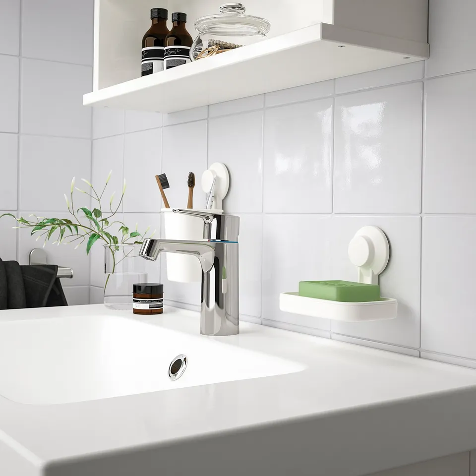 Modern white bathroom sink with chrome tap, small plant, toothbrushes, and green soap in white TISKEN holder.