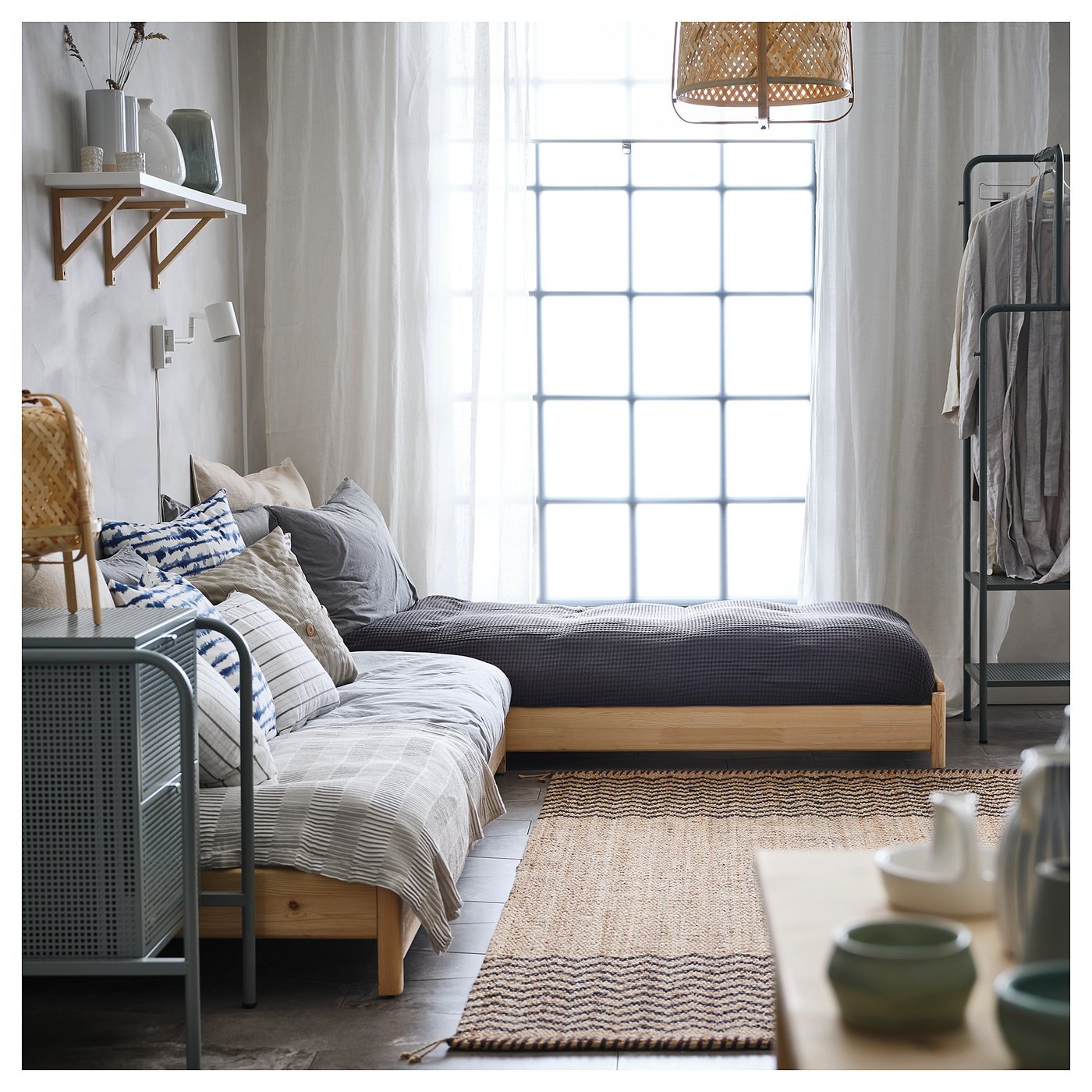 Sovereign minimalist living space featuring wooden UTÅKER bed with cushions against bright window, patterned rug, and wicker chair.