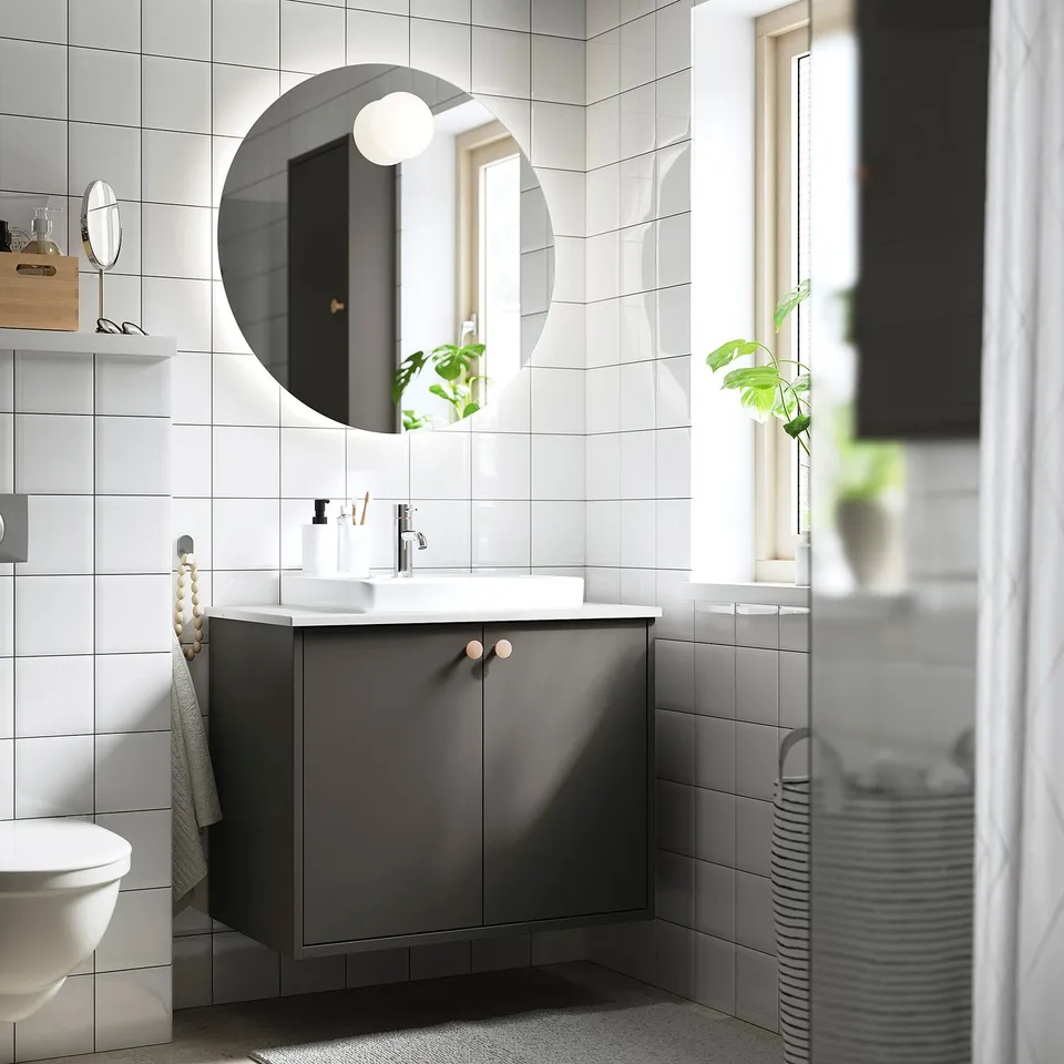 Modern minimalist bathroom: round mirror, white-tiled walls, grey HAVBÄCK vanity with sink, grey towel, plant, and striped laundry basket.