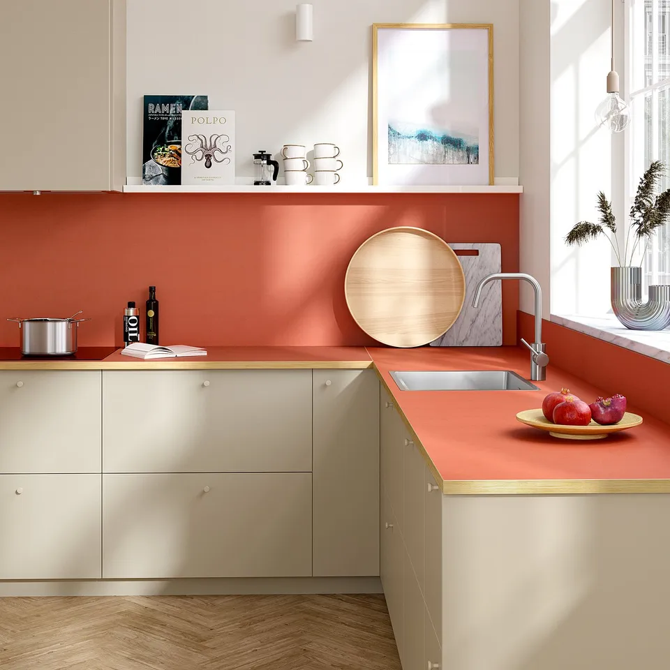Modern kitchen with red EKBACKEN countertop, sink, muted beige cabinets, bowl of pomegranates, utensils, and artwork.