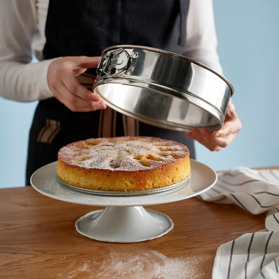 Person removing a round cake from a springform pan on a cake stand. The pan has a lock mechanism and is being used to bake a golden cake.