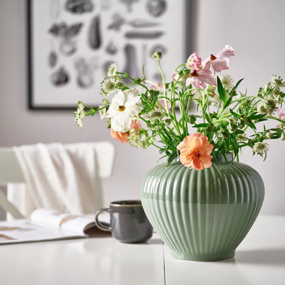 Green glazed stoneware vase holds colourful flowers on a table with a grey mug and a white chair nearby.