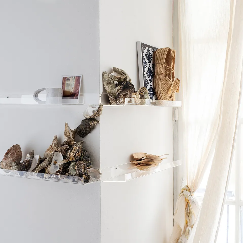 Three acrylic white shelves display oyster shells, framed art, woven baskets. Soft light from sheer curtains.