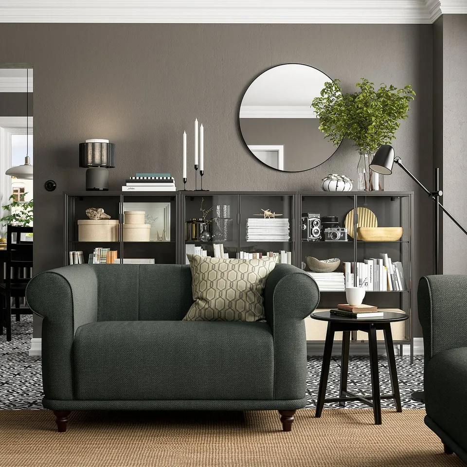 Cosy living room with dark walls, VISKAFORS green armchair, patterned rug, black bookshelf with plants, books, and a round mirror.