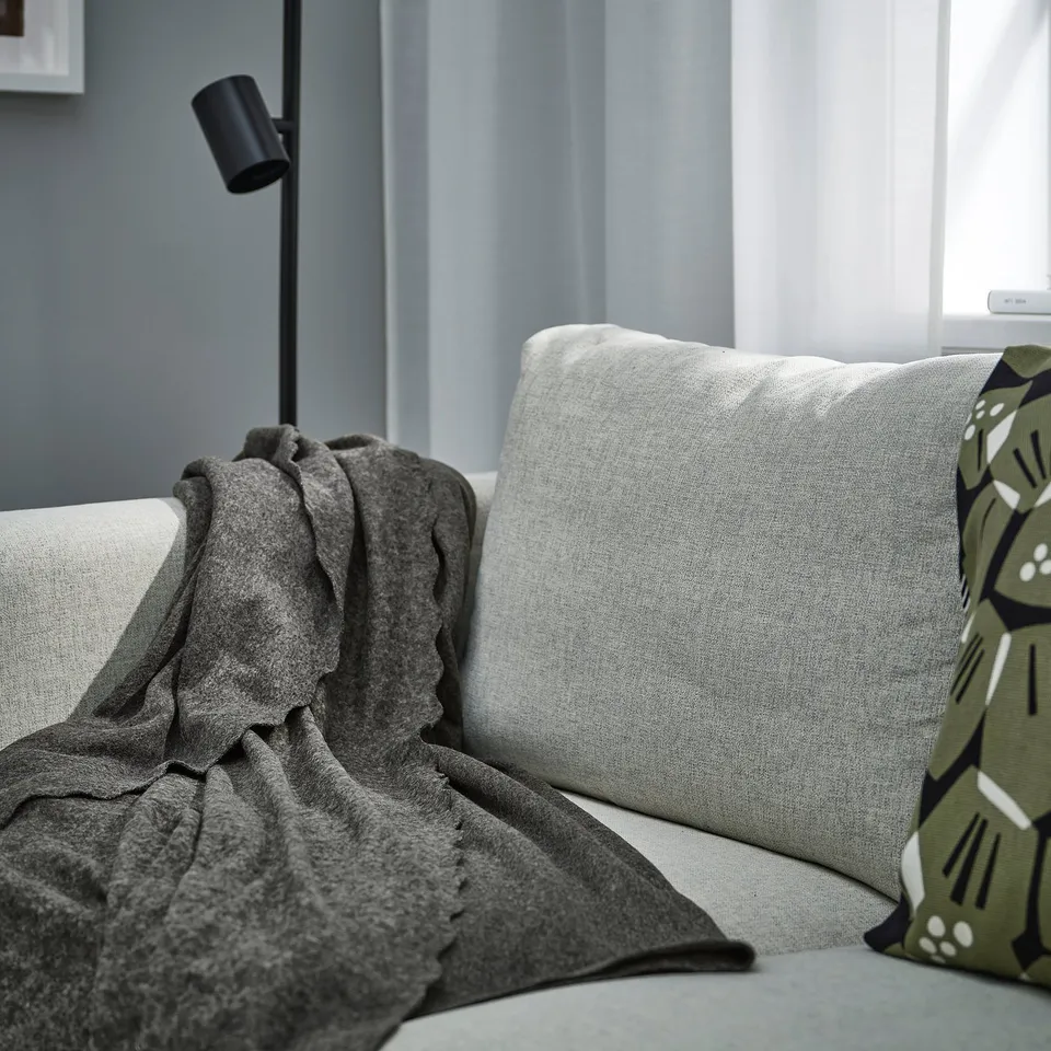Dark grey DAGGKÅPOR fleece throw draped on light sofa, partially covering large grey pillow, beside modern black lamp; cosy & recycled.