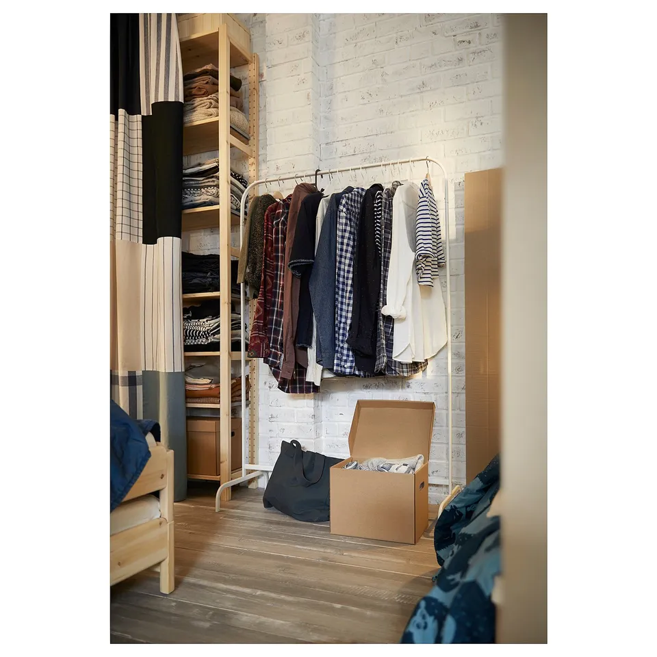 Minimalistic metal & plastic clothes rack with assorted clothes against white brick wall, plus floor box & bag.