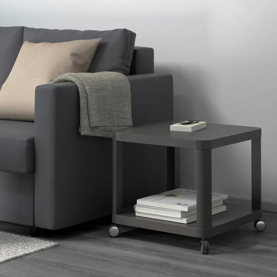 Dark grey sofa with light-eye cushion, grey throw over arm. Beside it, black rolling side table with books and remote on top.