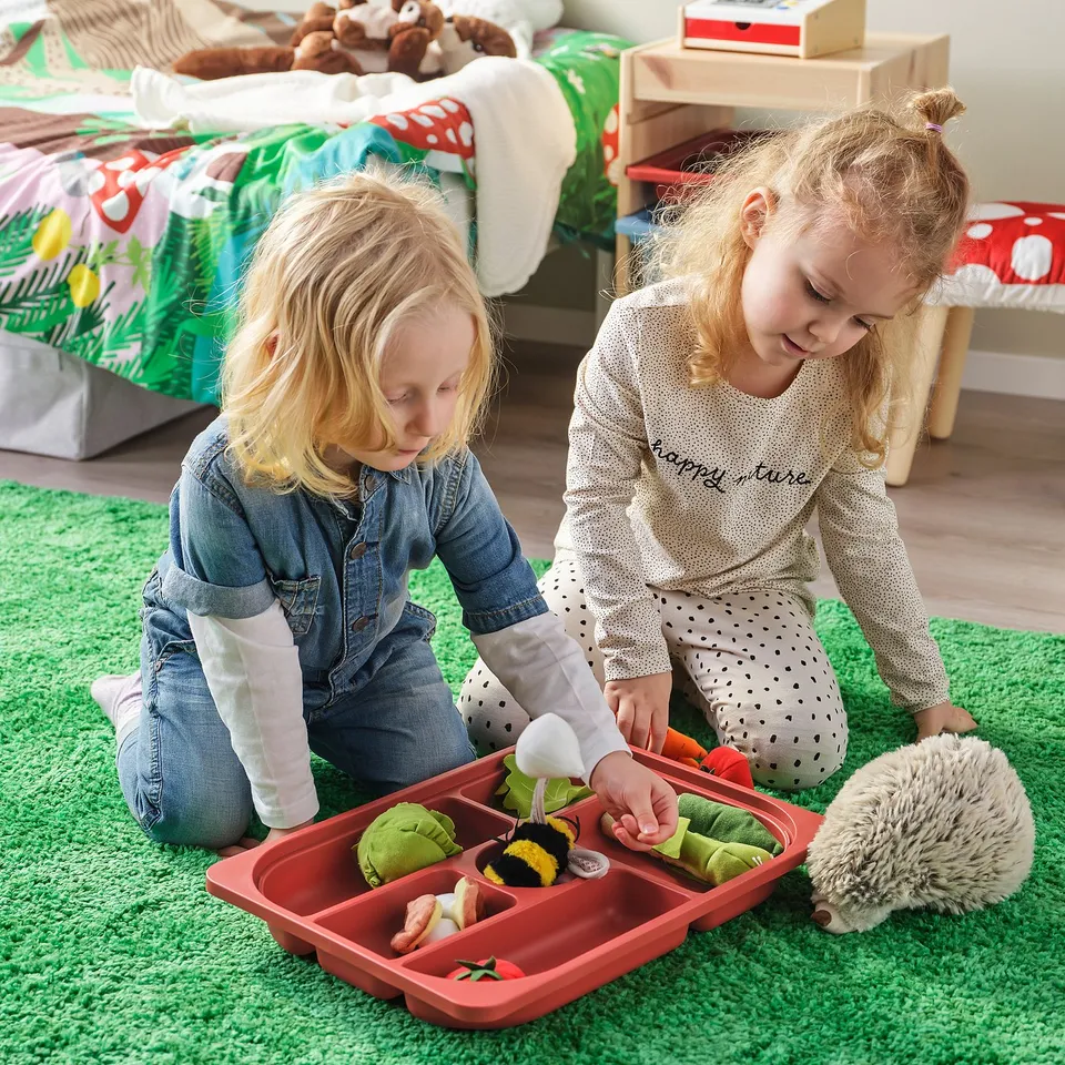 Two children play on a green rug with a red TROFAST tray full of stuffed animals and other toys, organising and enjoying their playtime.