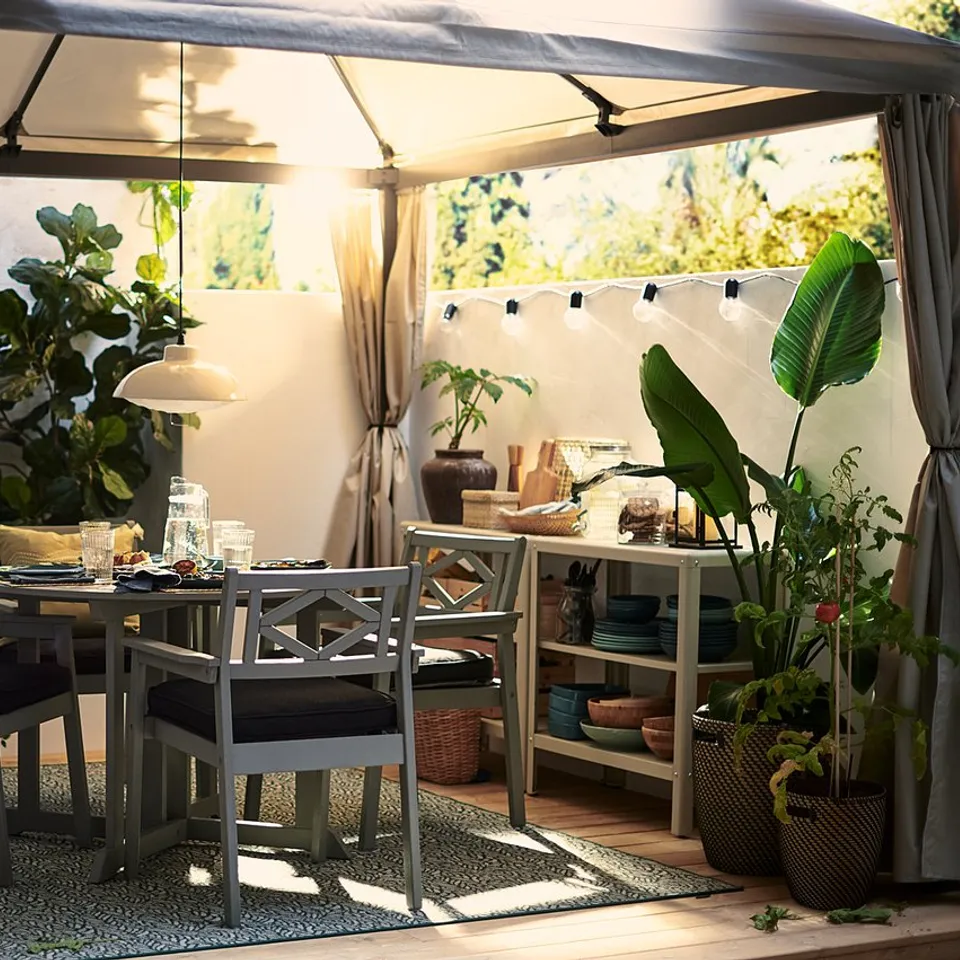 Outdoor dining setup with grey himmelso gazebo, wooden table, black chairs, plants, and string lights.