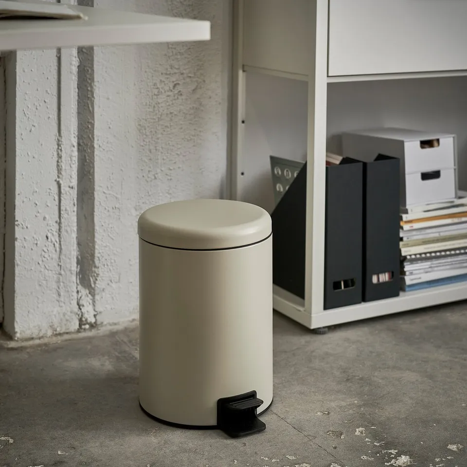 Small step bin in a beige colour placed on a concrete floor next to a white shelf with folders.