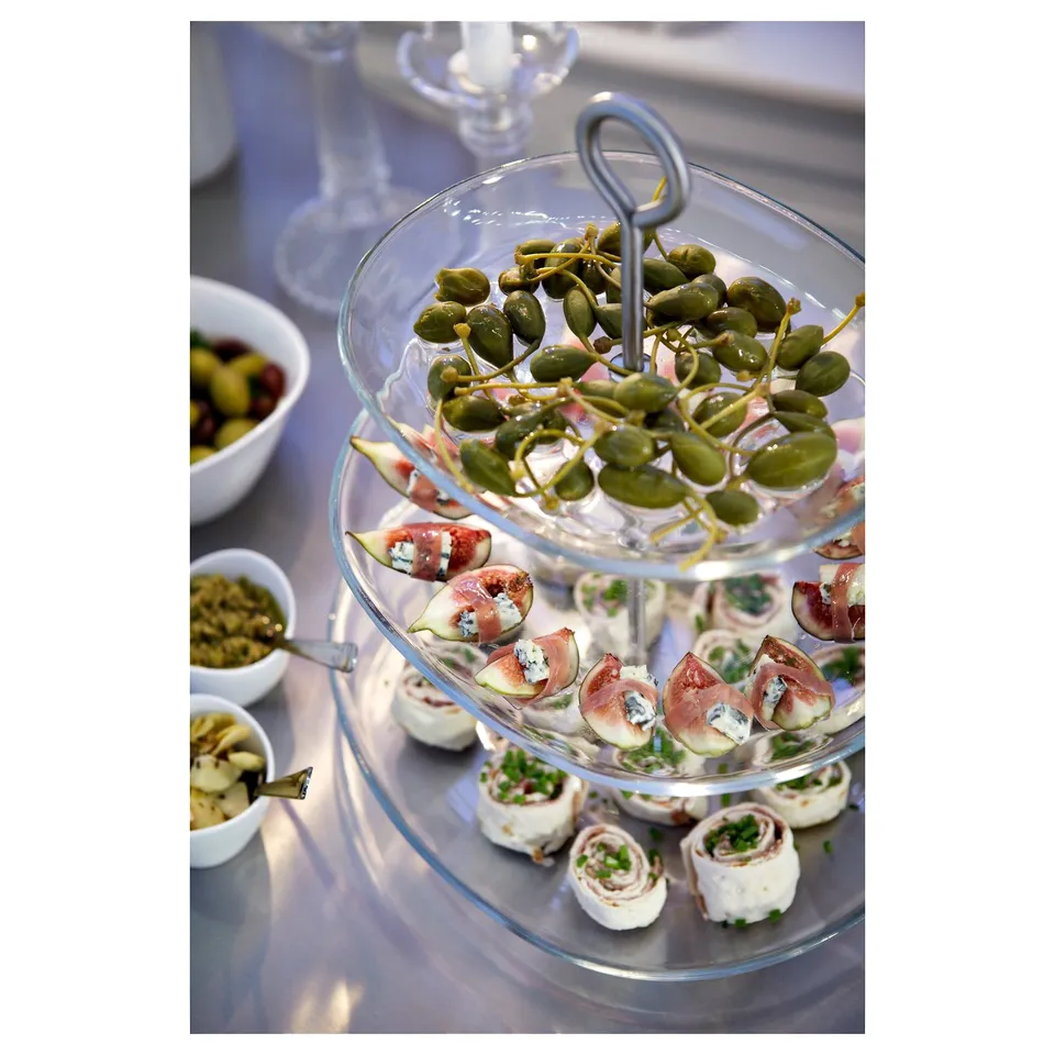 Clear glass tiered food stand holding appetisers, featuring caprese skewers and wrapped snacks. Elegant and modern kitchen accessory.