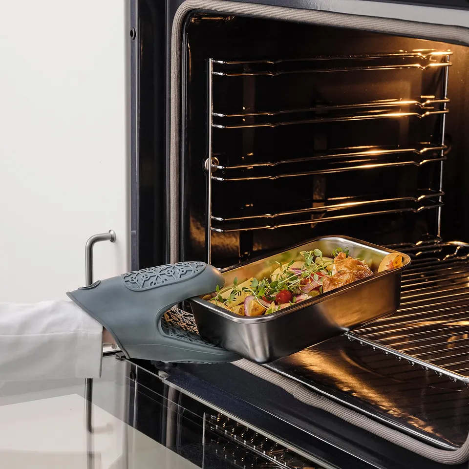 Open oven with stainless steel pan filled with roasted vegetables and meat, placed on a silicone mitt.