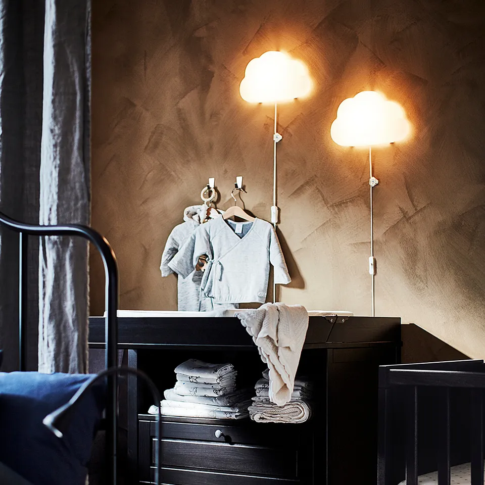 Cosy nursery with black furniture, UPPLYST lamps, and soft light. Grey clothes, towels, and a dark blue blanket decorate the wooden space.