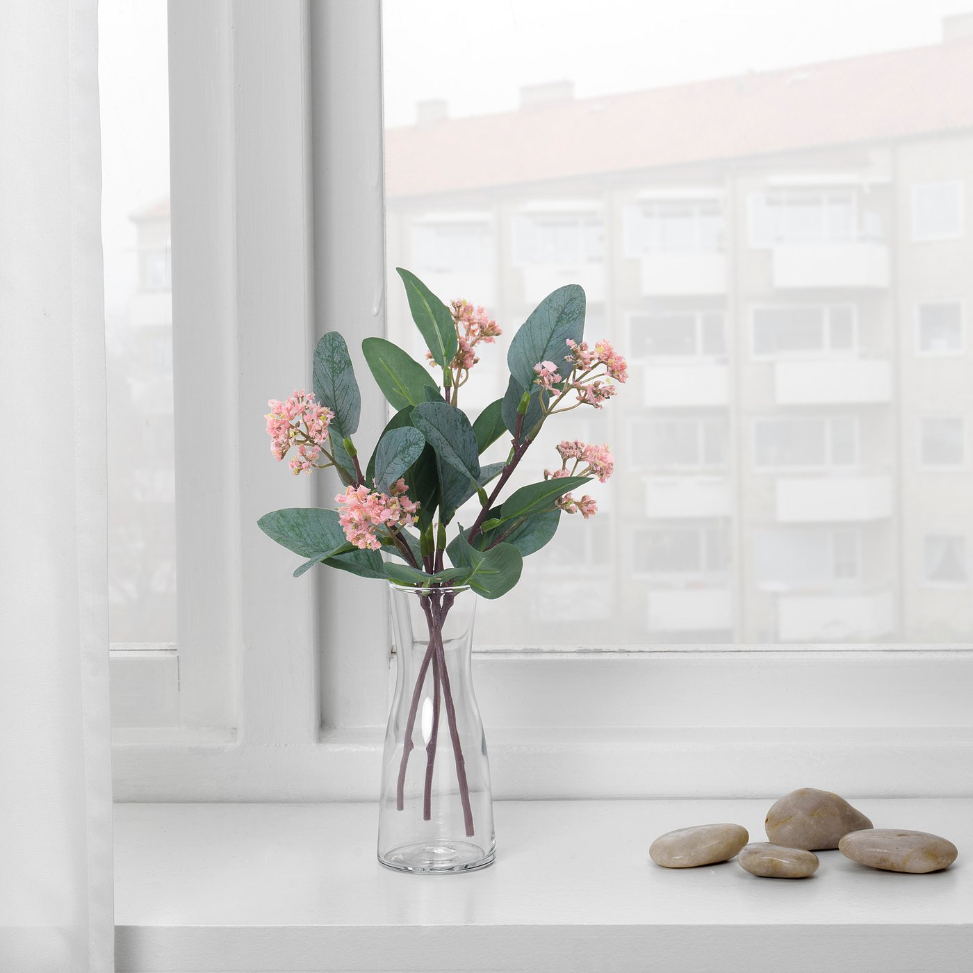 A clear vase with pink artificial flowers sits on a white windowsill, showcasing a fresh, low-maintenance, and eco-friendly decor.