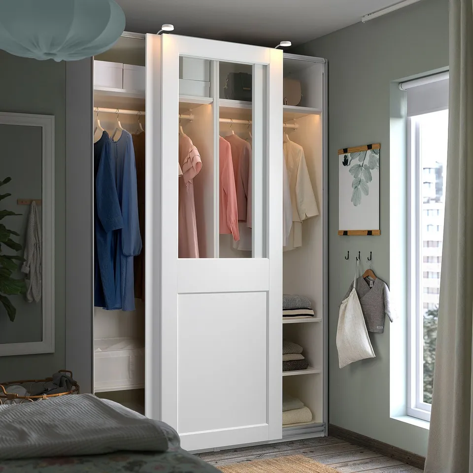 Modern bedroom with a tall white PAX wardrobe featuring glass doors, well-organized with open and closed storage near a window.