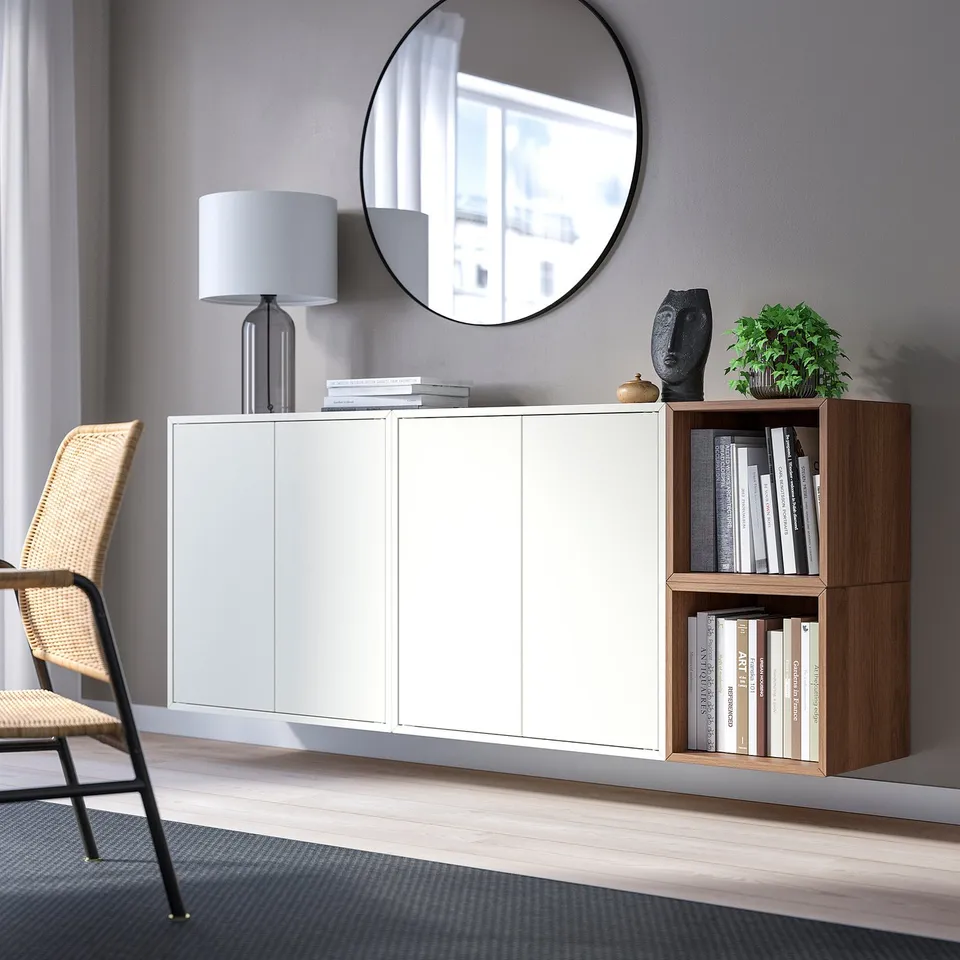 Modern living room with EKET cabinets, wooden shelves, and a round mirror. Minimalist design with books, lamp, and wicker chair.