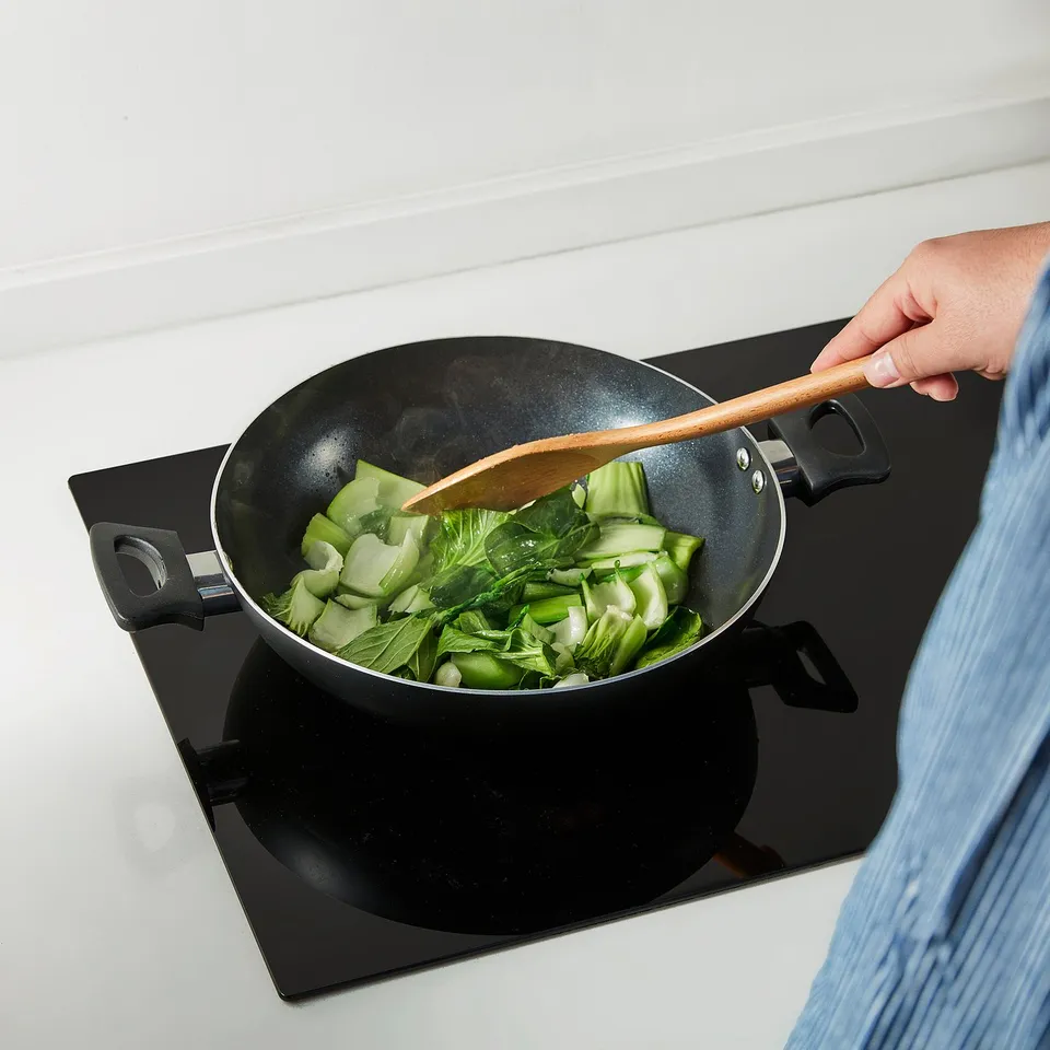 A person stir-fries vegetables in a two-handled non-stick kadai wok using a wooden spatula.