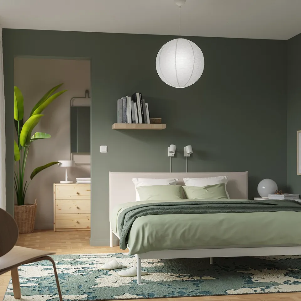Modern bedroom with white GULLSUDARE lamp above bed, green bedding, wooden furniture, plant, and patterned rug.