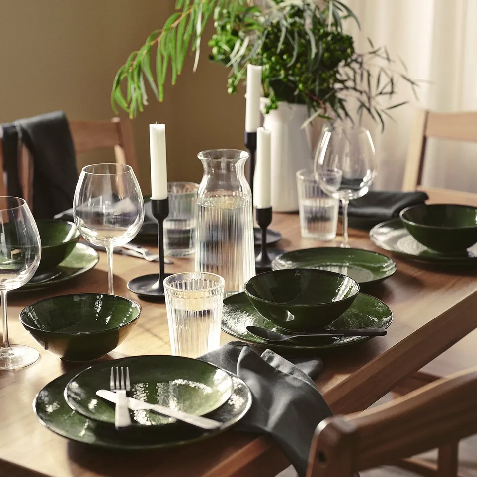 Green PELARKAKTUS dinnerware, plates, bowls, glasses, cutlery, napkins, candles, and greenery vase on table.