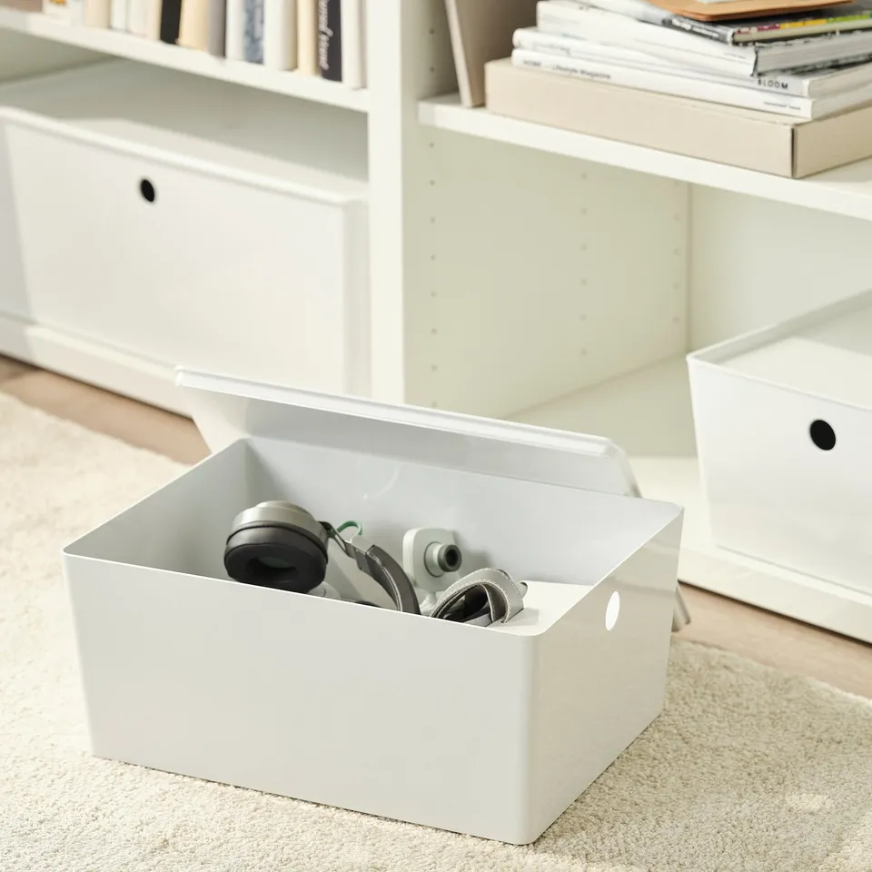 White storage box with lid on carpet, holding rolled items. Part of KUGGIS series, useful for organising gadgets or papers.