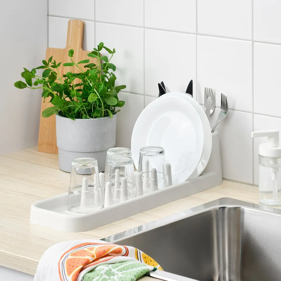 Kitchen sink with white dish drainer holding plates, glasses, and cutlery. A potted plant, soap dispenser, and colourful dish towels are also present.