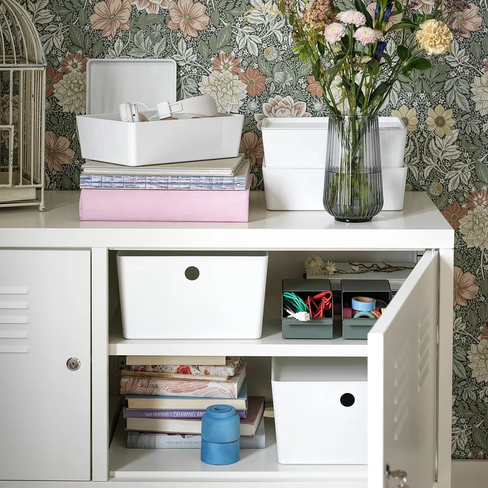 White IKEA PS metal cabinet with storage boxes, books, and flowers. Floral wallpaper background.