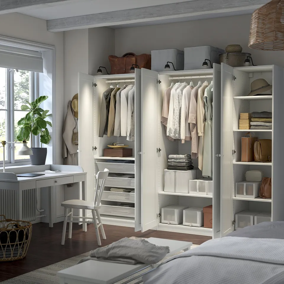 Well-organized bedroom with white PAX wardrobe units displaying clothes, shelves with accessories, and a desk with a plant beside a window.
