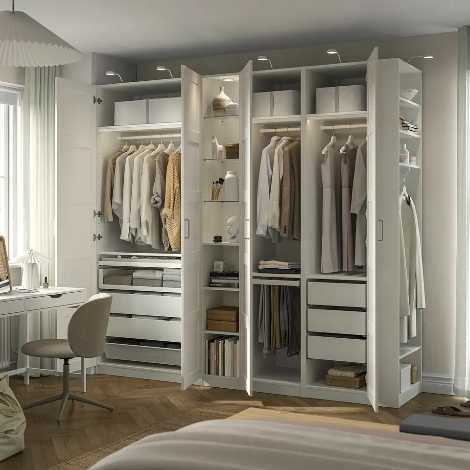 Modern bedroom with PAX wardrobe, open doors showing organised clothes. Integrated lighting, desk and chair nearby.