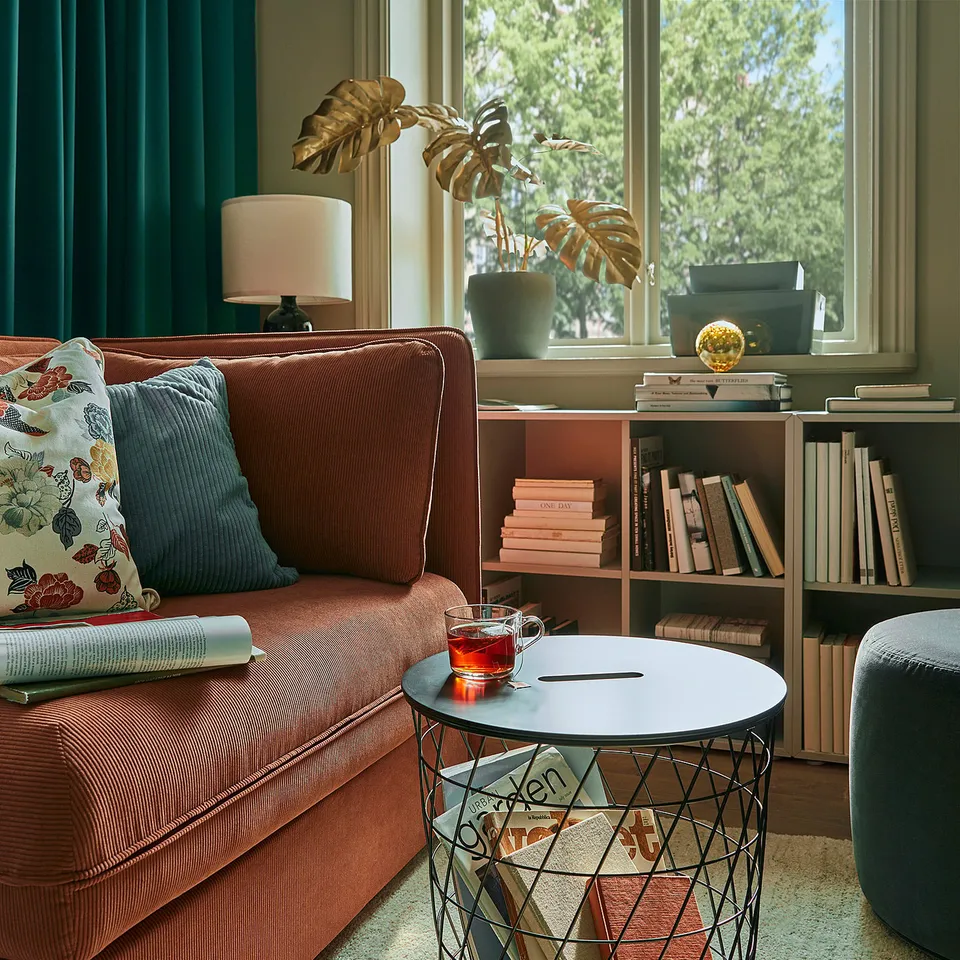KVISTBRO red side table with books and cup, brown cushions, blue curtains, near grey rug.