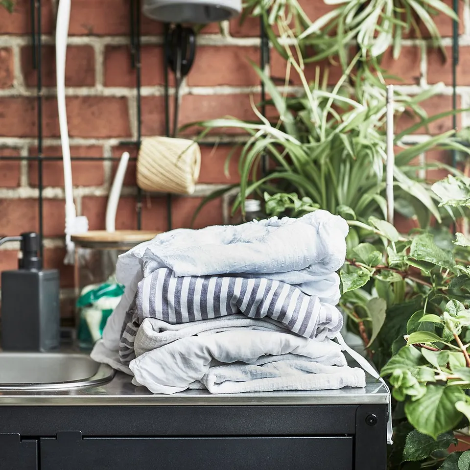 Stack of folded blue striped towels on black outdoor sink shelf, brick wall background, surrounded by green plants.