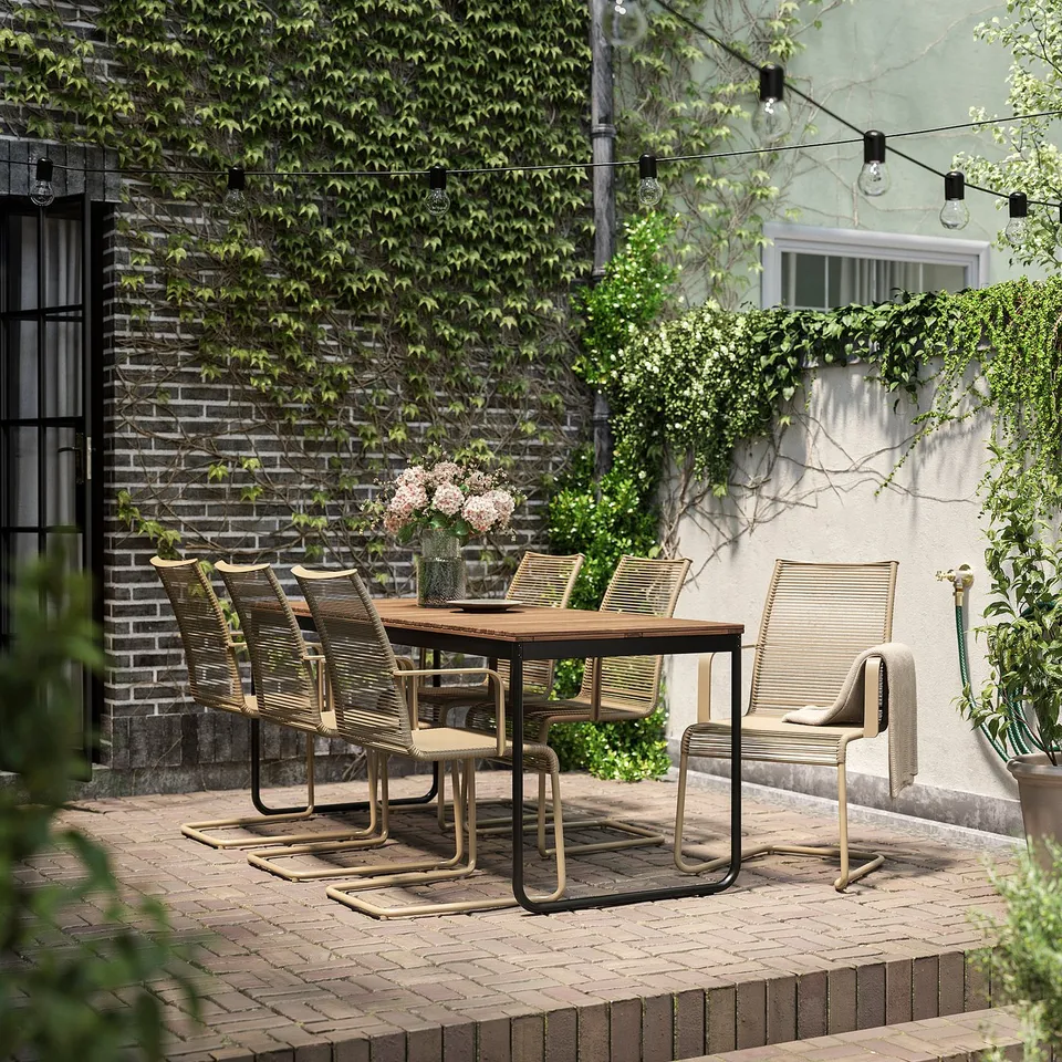 Outdoor dining set with wood table and wicker chairs, surrounded by greenery and string lights.