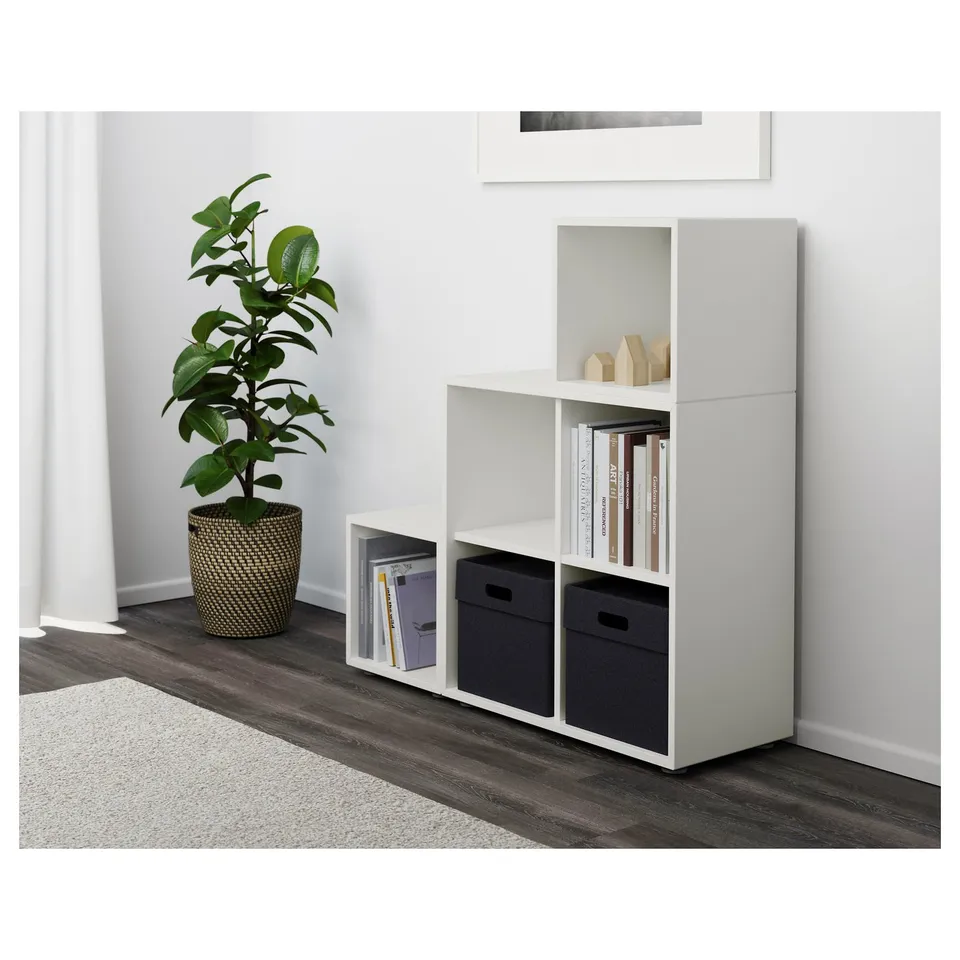 Modern white shelving unit with adjustable storage, holding books and black boxes, next to a tall green plant.
