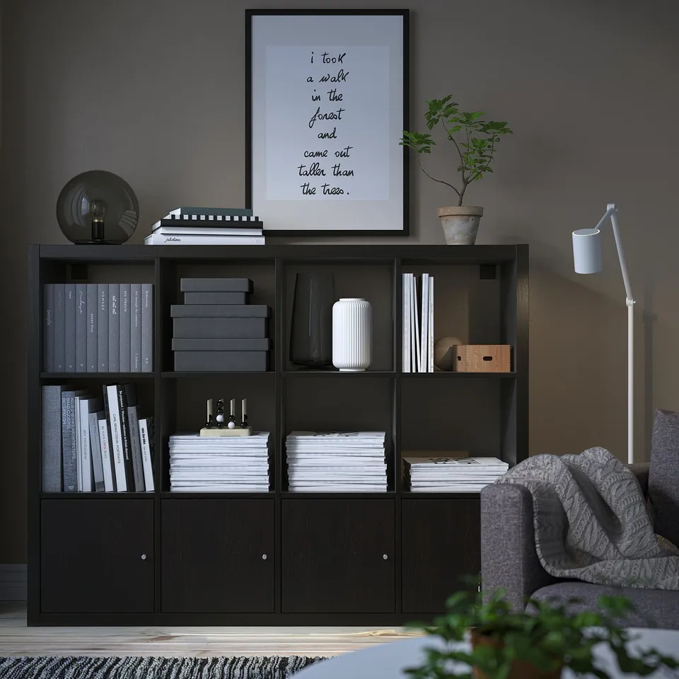 Black KALLAX bookshelf filled with books and decorations, against dark wall, next to grey couch, with frame and plant on top.