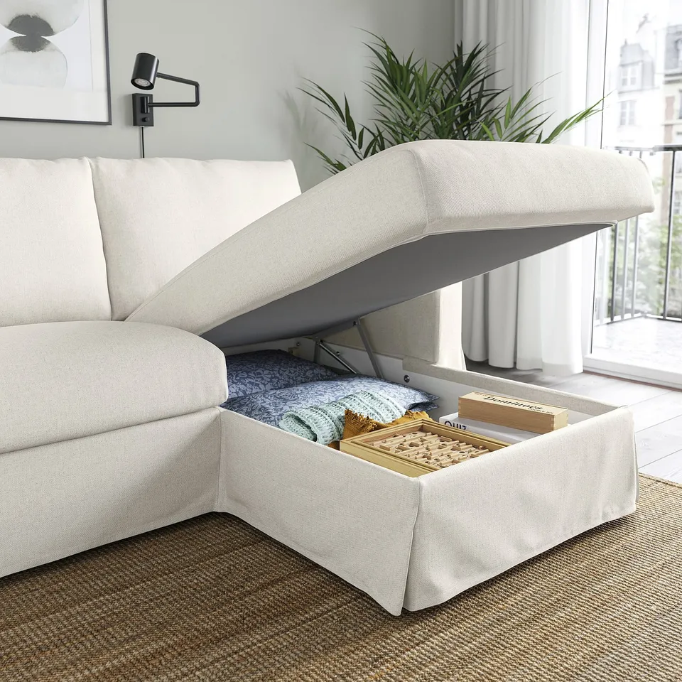 White sectional sofa with open ottoman storage, revealing blankets and boards, near a window with plants.