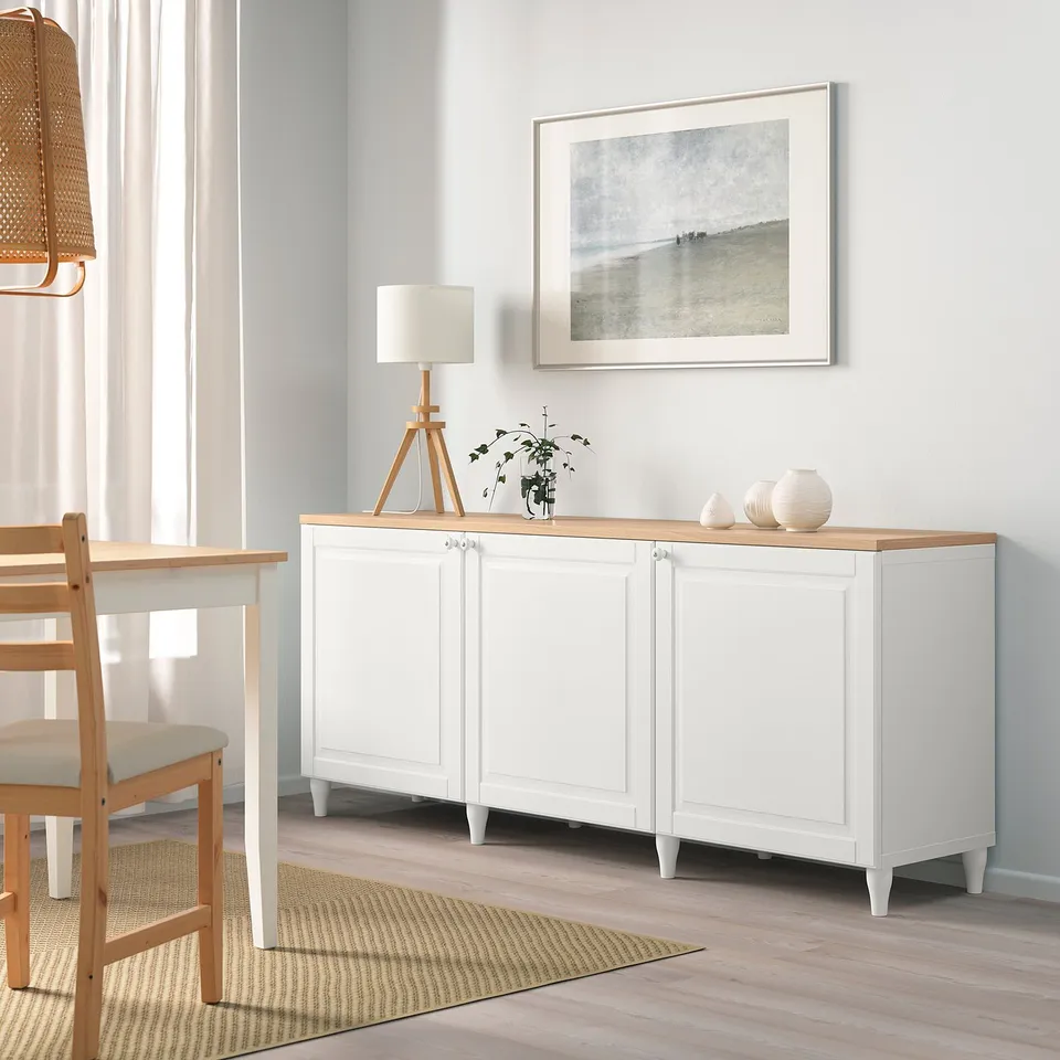 White BESTÅ sideboard with oak top. Contains decor, lamp, plant, and framed art. Large in size compared to chairs.