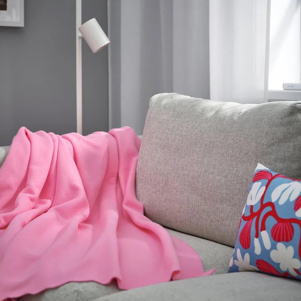 Cosy pink DAGGKÅPOR fleece blanket on light grey couch. Soft, recycled polyester. Large, draped over armrest, near colourful pillow.