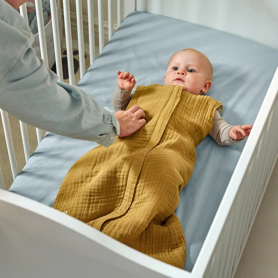 Baby in mustard sleep bag, being zipped up by adult. Light blue crib, light wood floor.