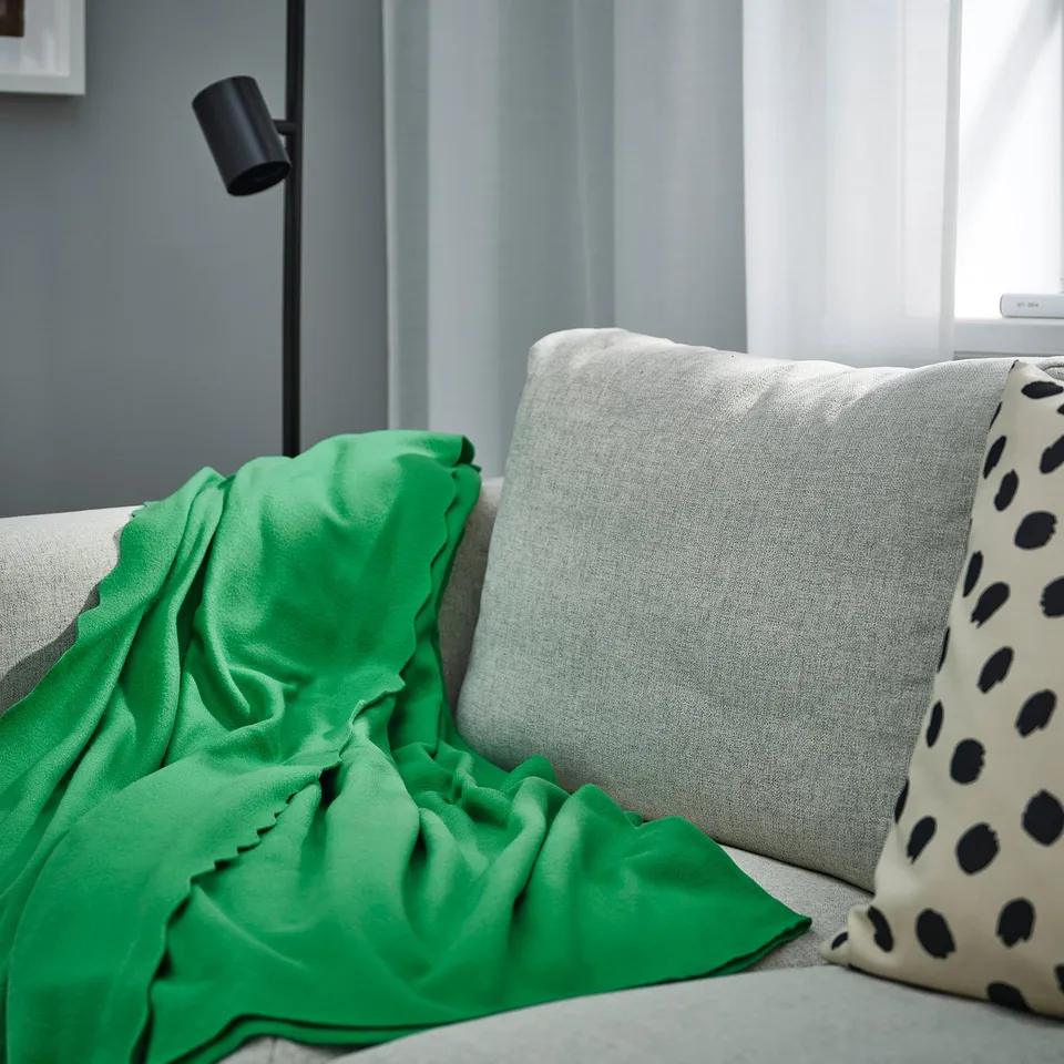 Green DAGGKÅPOR fleece blanket draped on large grey couch with spotted pillow, near black lamp.