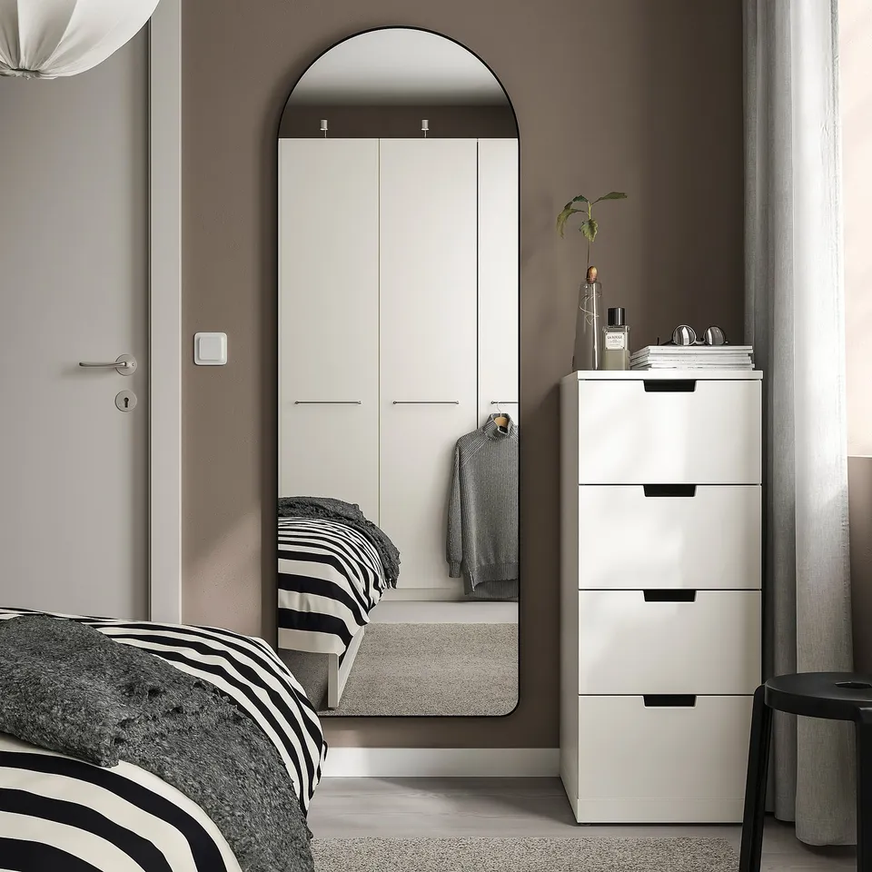 Modern bedroom with arched LINDBYN mirror, reflecting striped bedding. White dresser with black handles, grey stool, neutral walls.