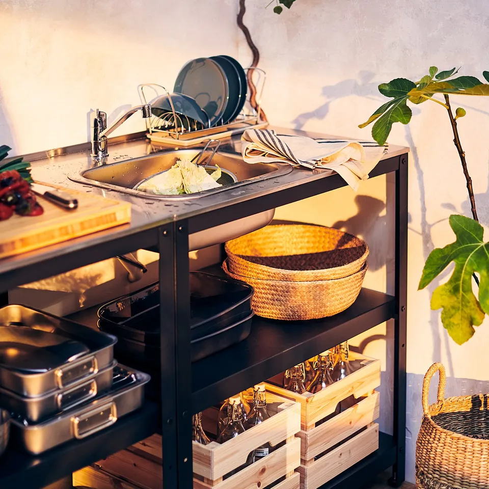 Black GRILLSKÄR outdoor sink with shelves. Stainless steel top, wicker baskets, cooking tools, potted plant nearby.