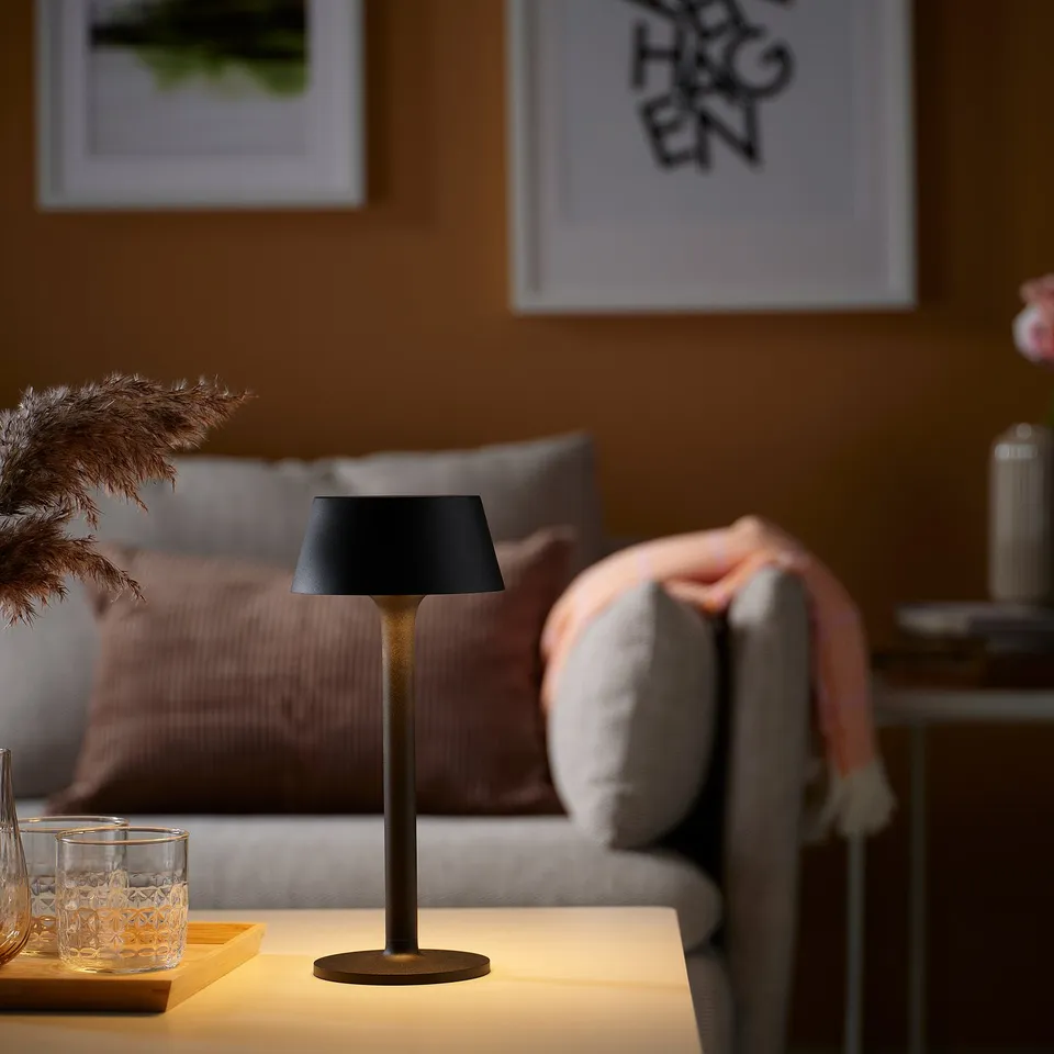 Small black AVHÅLL lamp on table near couch, with drinks. Cosy indoor lighting.