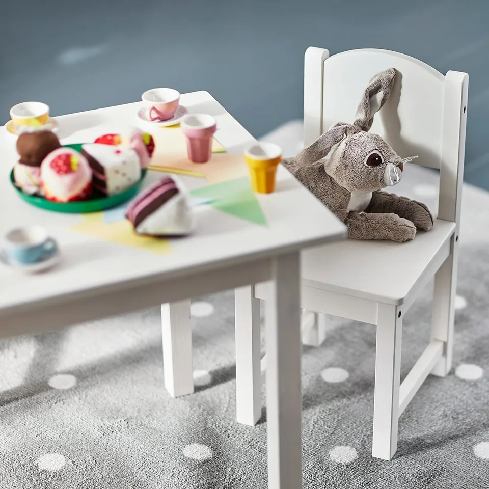 Small childs white wooden chair with grey stuffed animal sitting, next to play table with colourful toys and cups.
