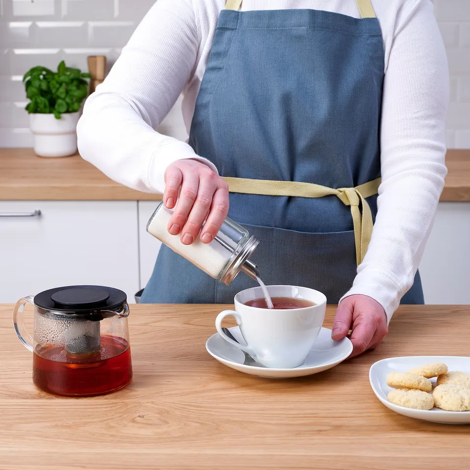 Person wearing an apron pours tea into a cup from a glass container. A teapot with infuser and cookies are on the table.