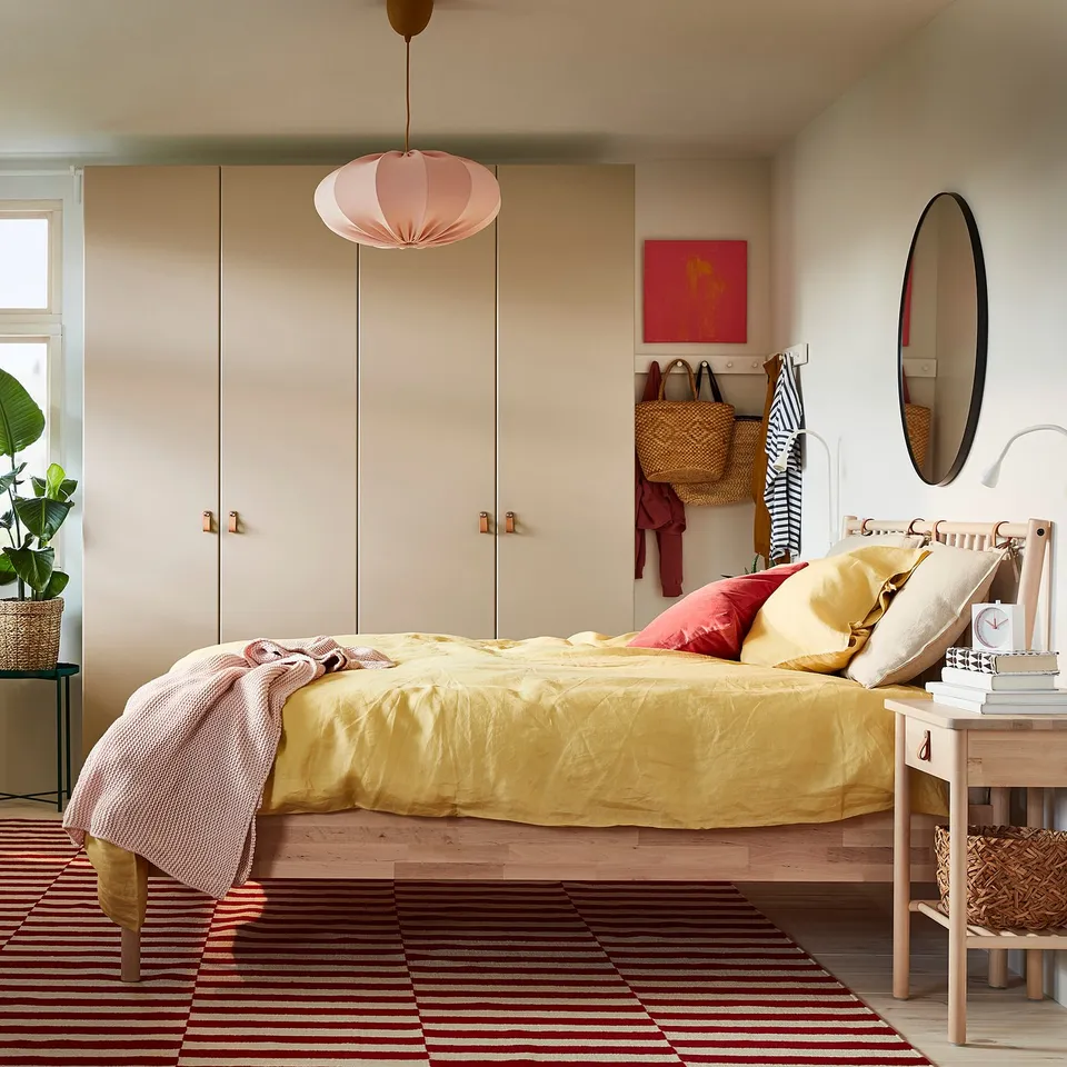 Bright, cosy bedroom with yellow BJÖRKSNÄS bed, striped rug, pink pendant light, and round mirror. Natural wood floors and beige walls.